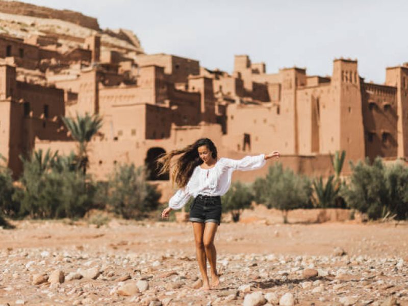 Young curly woman eastern appearance walking on background of kasbah Ait-Ben-Haddou. Travel in Morocco, Ouarzazate. Summer vacations, travel lifestyle concept.